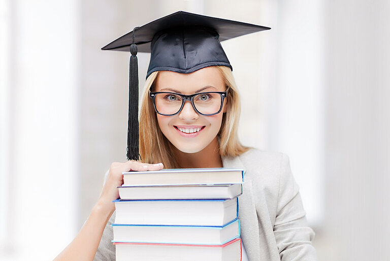 Mädchen hat einen Doktorhut auf, lacht und hat vor sich einen Stapel Bücher